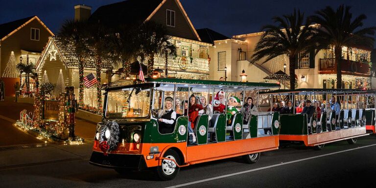 A festive green and orange trolley decorated with holiday wreaths driving through glowing white lights in St. Augustine.
